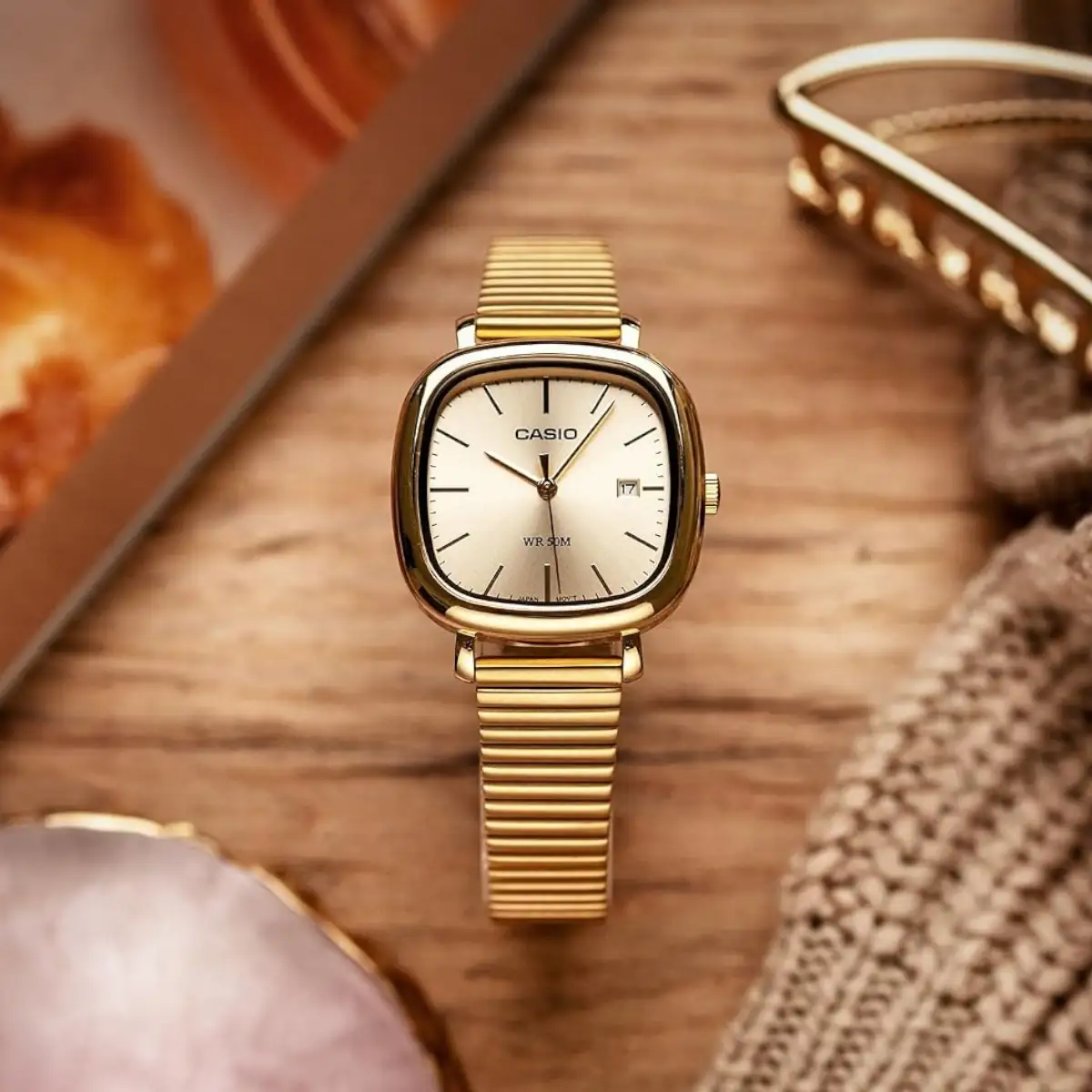 #image_title Gold Casio wristwatch with a square rounded face and a metal expansion band resting on a wooden surface, showing a small date window.
