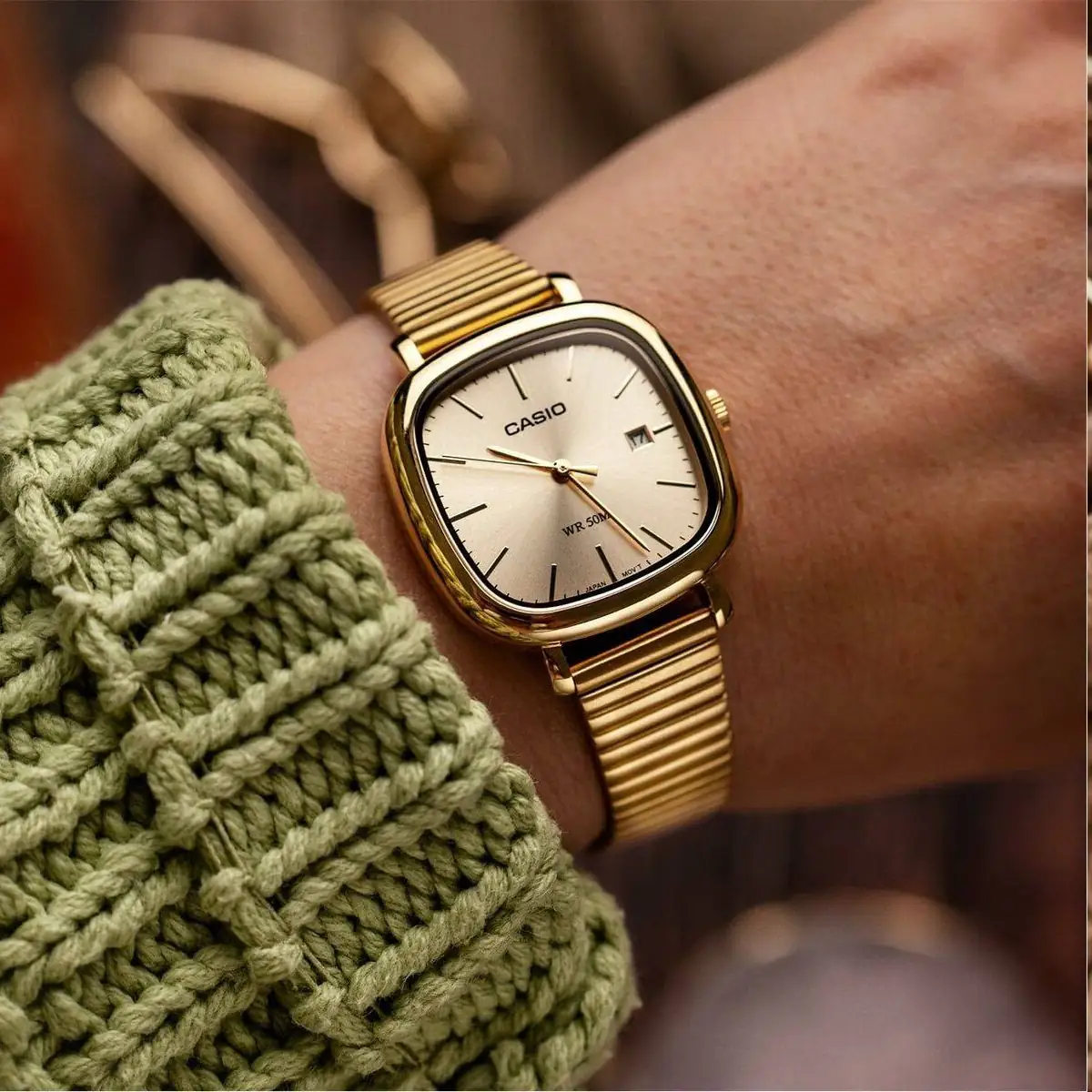 #image_title Gold Casio wristwatch with a cream dial and date window, worn over a green knitted sleeve/ sweater.